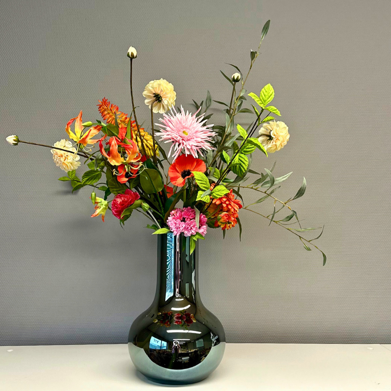 Boeket Aayushi in de elegante, spiegelende Vaas Seim met kleurrijke zijden bloemen zoals gloriosa lelies, dahlia's, klaprozen en anjers, gepresenteerd op een lichte achtergrond