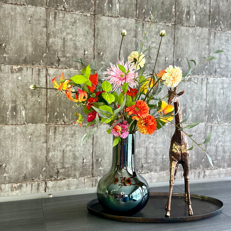 Boeket Aayushi in de elegante, spiegelende Vaas Seim met kleurrijke zijden bloemen zoals gloriosa lelies, dahlia's, klaprozen en anjers, gepresenteerd op een decoratieve schaal en bronzen giraffe